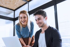 Dos jóvenes profesionales colaboran sonrientes frente a un portátil en una oficina luminosa, reflejando las ventajas de ser funcionario TIC: trabajo en equipo y flexibilidad horaria.