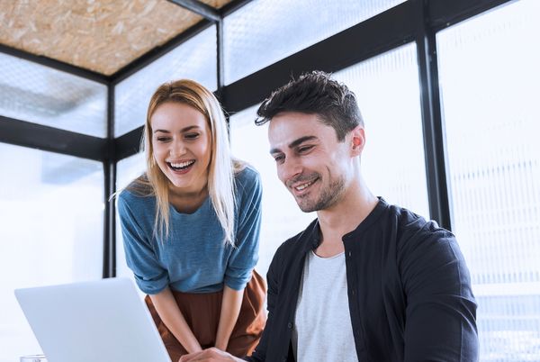 Dos jóvenes profesionales colaboran sonrientes frente a un portátil en una oficina luminosa, reflejando las ventajas de ser funcionario TIC: trabajo en equipo y flexibilidad horaria.