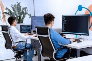 Dos opositores TIC trabajando frente a ordenadores con pantallas de código, representando los distintos perfiles según el tipo de oposición TIC que elijan.
