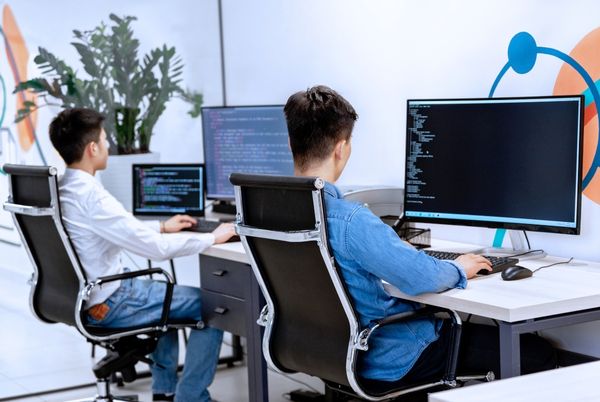 Dos opositores TIC trabajando frente a ordenadores con pantallas de código, representando los distintos perfiles según el tipo de oposición TIC que elijan.