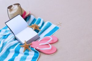 Toalla en la playa con libro, chanclas y sombrero, símbolo de estudiar oposiciones TIC en vacaciones.