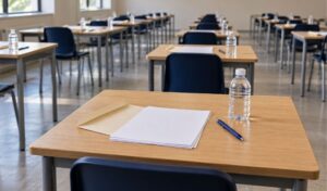 Aula de examen vacía con mesas individuales, hojas en blanco, sobre abierto, bolígrafo azul y botella de agua sin etiqueta.